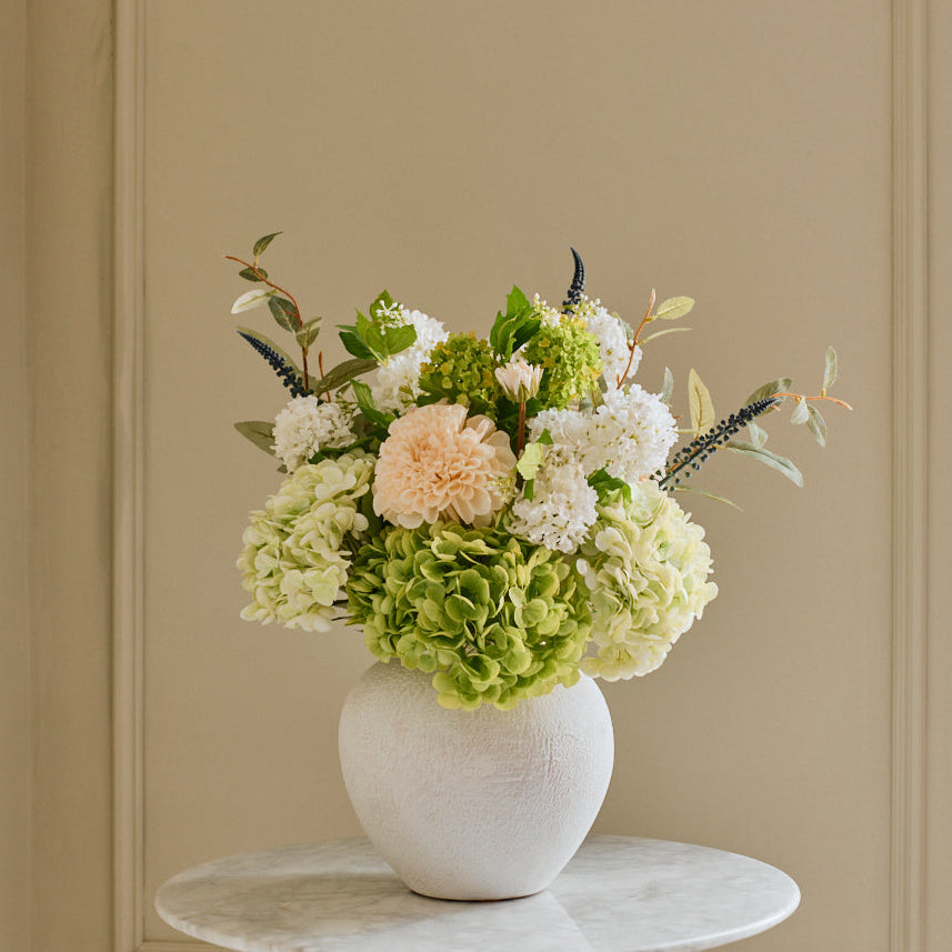 Floral arrangement in a white vase on a small round table against a beige paneled wall.