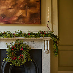 A holiday wreath made of green pine branches and red berries hangs on a black fireplace surround, with a matching green garland draped across a white mantel. A framed abstract painting and a candle are visible in the room.