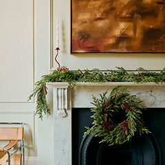 A holiday-themed interior scene featuring a white marble fireplace mantel decorated with a green garland and a green wreath with red berries, beneath a large abstract brown painting.