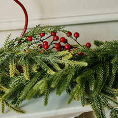 A green pine garland with clusters of red berries draped across a white mantel, with a red candlestick visible in the background against a cream wall.