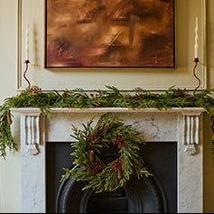 A holiday wreath made of green pine branches and red berries hangs on a black fireplace surround, with a matching green garland draped across a white mantel. A framed abstract painting and a candle are visible in the room.