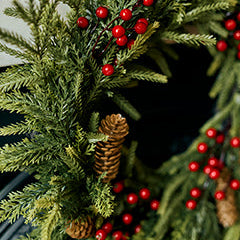 A close-up of a green evergreen wreath decorated with small red berries and natural pinecones.