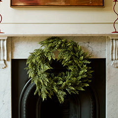 Conifer Cypress Faux Christmas Wreath. A green evergreen wreath hanging on a white marble fireplace mantel, with a large abstract brown painting displayed above it.