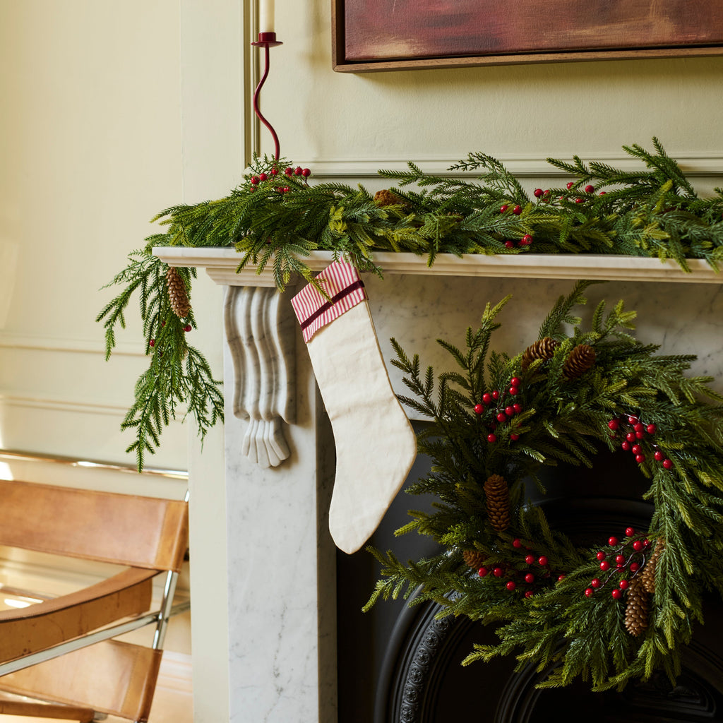A festive Christmas scene featuring a white striped stocking hanging from a white marble fireplace mantel decorated with a green pine garland and red berries, with a matching wreath on the black fireplace below.