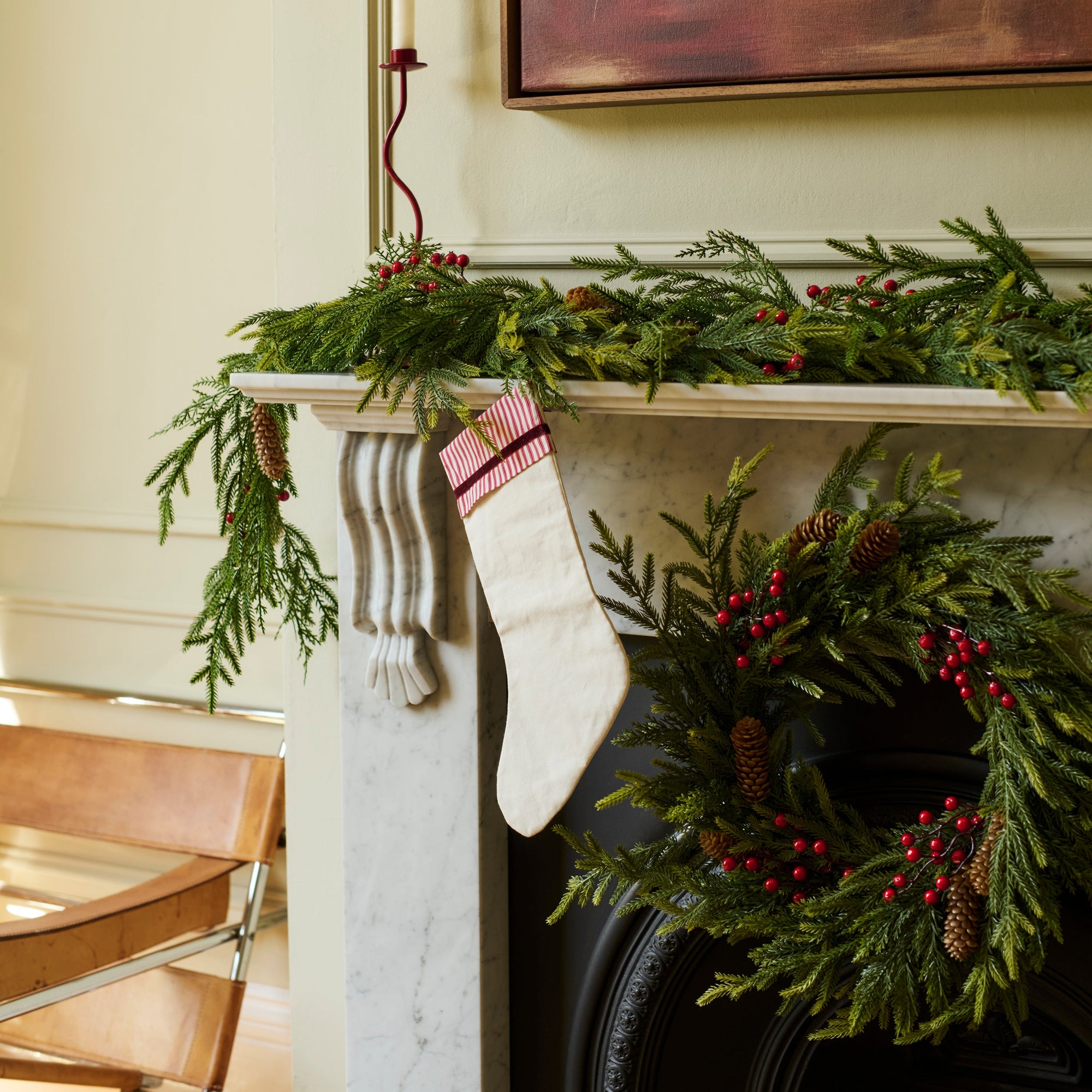 A festive Christmas scene featuring a white striped stocking hanging from a white marble fireplace mantel decorated with a green pine garland and red berries, with a matching wreath on the black fireplace below.