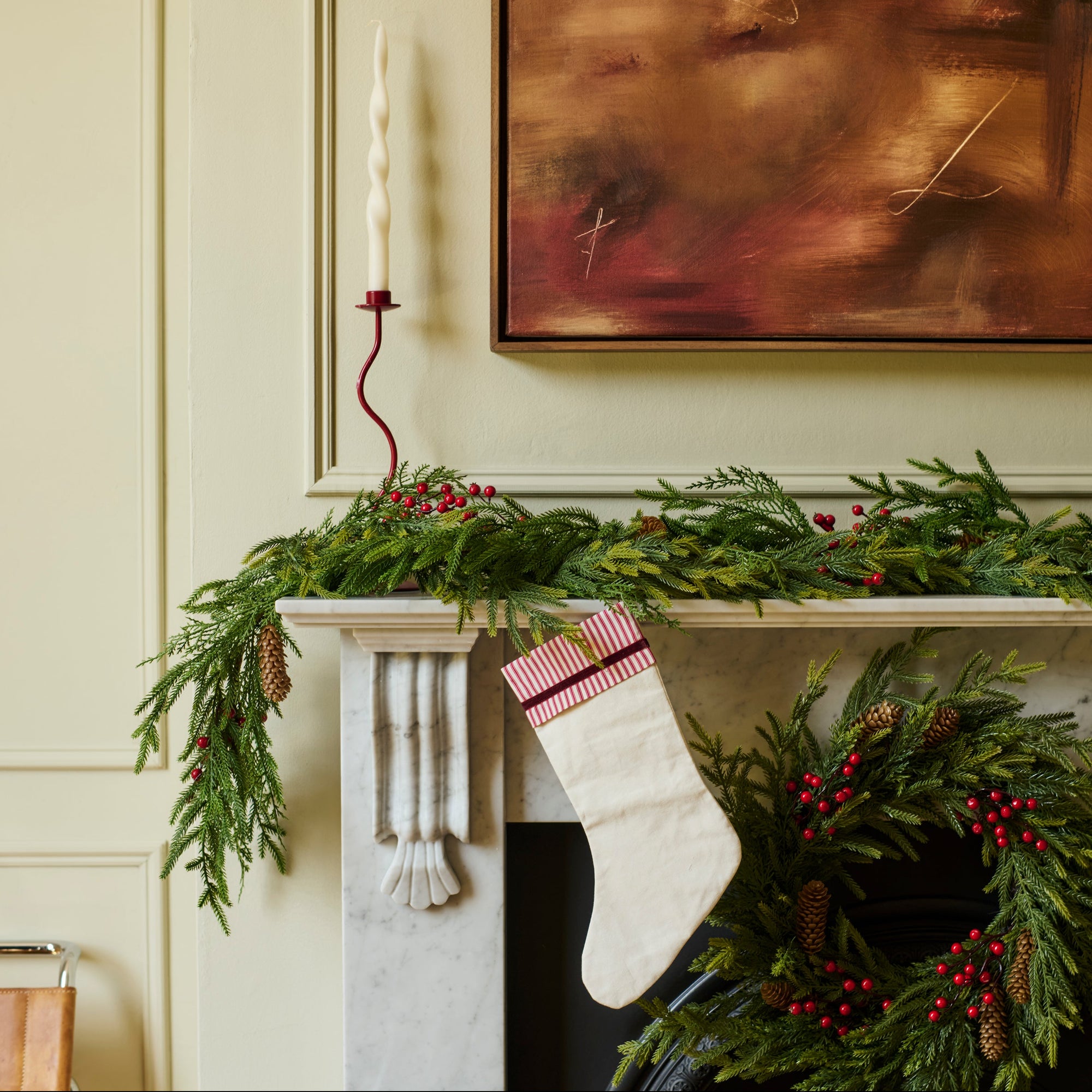 A festive Christmas stocking with a red and white striped cuff hangs from a white marble fireplace mantel decorated with a green pine garland and red berries. A matching pine wreath with red berries is draped over the fireplace opening below, set against a cream paneled wall with a framed abstract painting above.