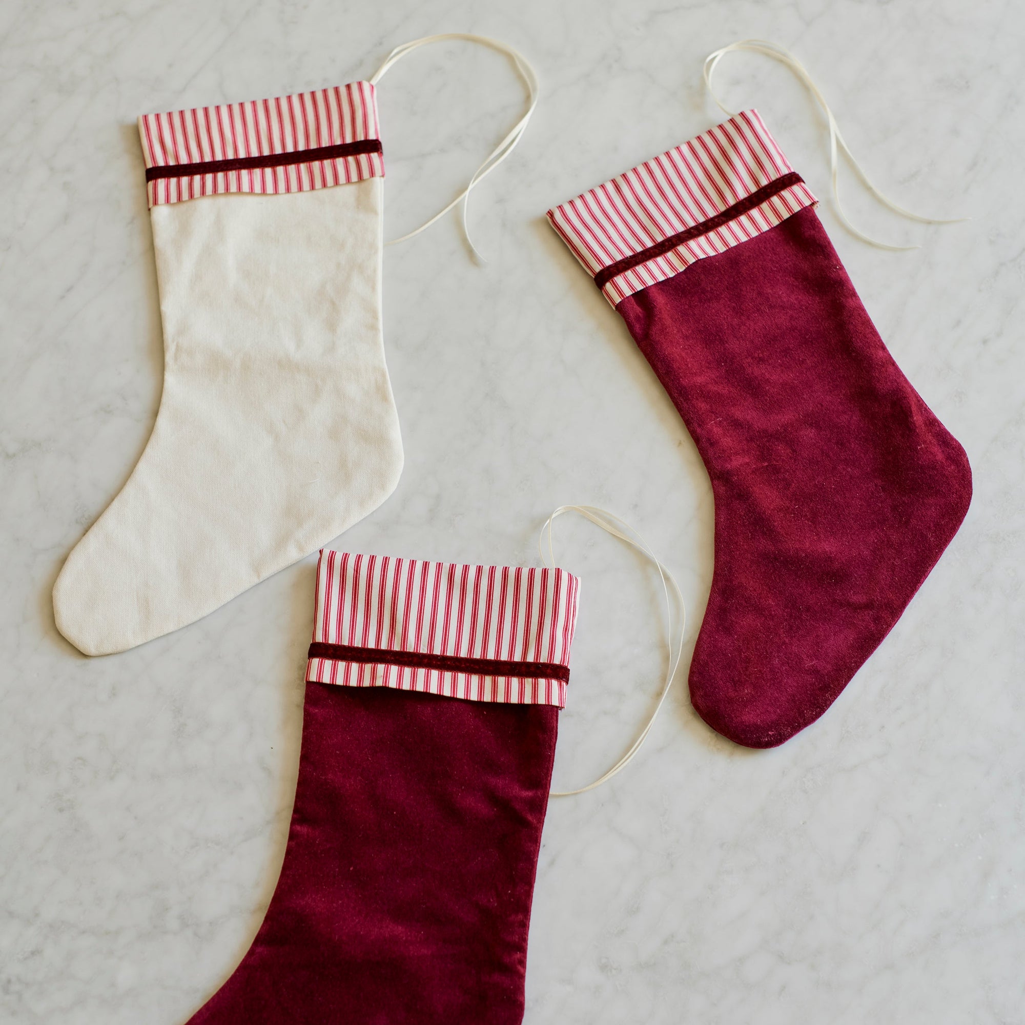 Three Christmas stockings laid out on a light marble surface: one cream stocking and two burgundy velvet stockings, each with a striped cuff and hanging loop.