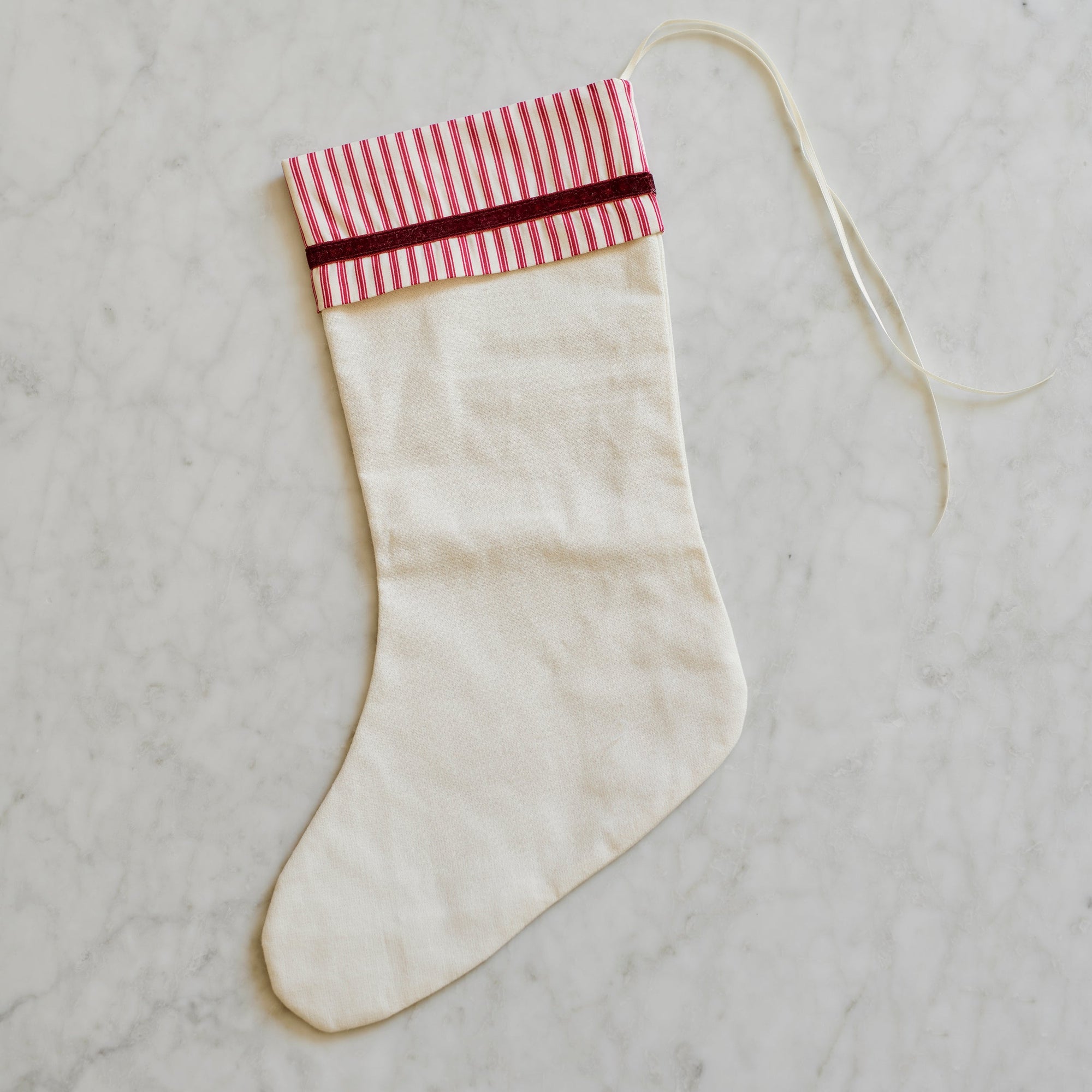 An off-white fabric Christmas stocking with a red and white striped cuff and a thin red stripe, laid flat on a light gray marble surface.