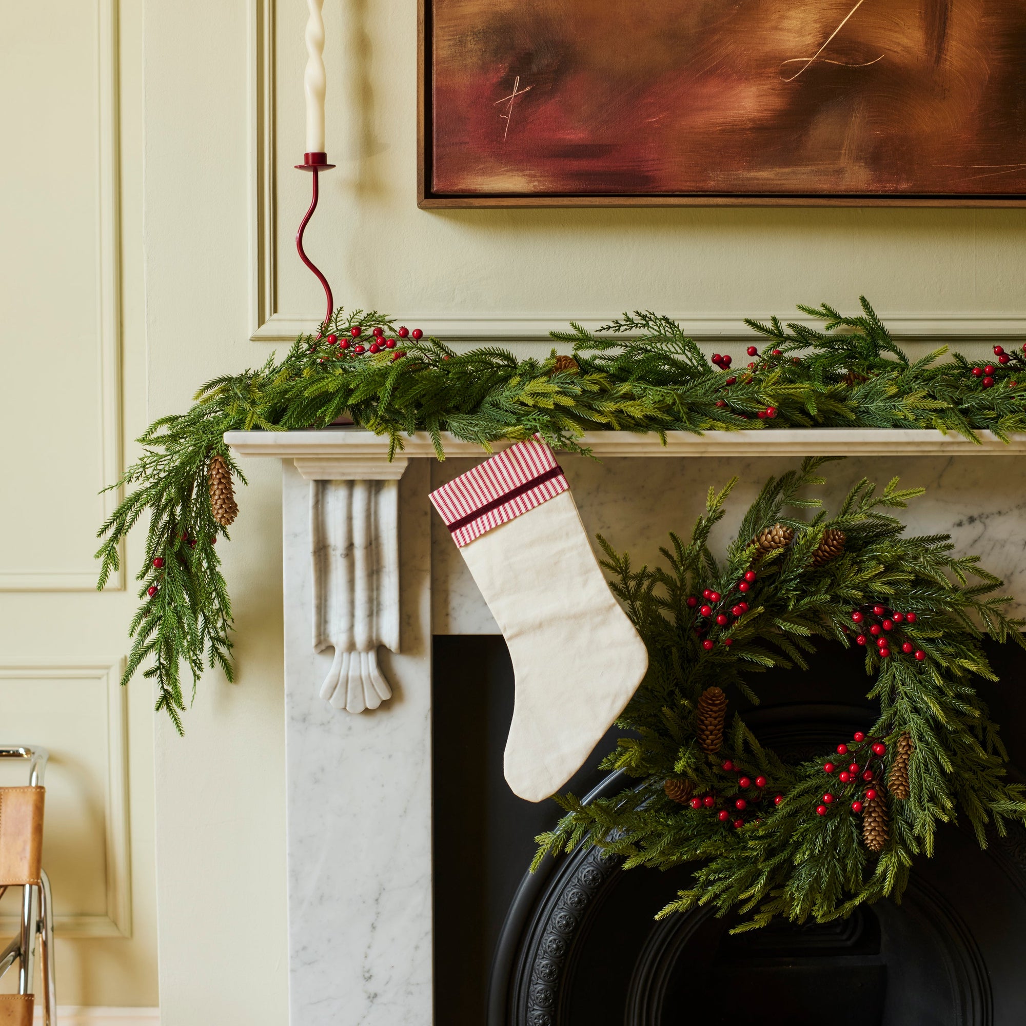 A festive holiday fireplace scene with a green garland draped across a white mantel, a white Christmas stocking with pink trim hanging from the left side, and a green wreath with red berries hanging on the right. A large abstract painting is mounted on the wall above the fireplace.