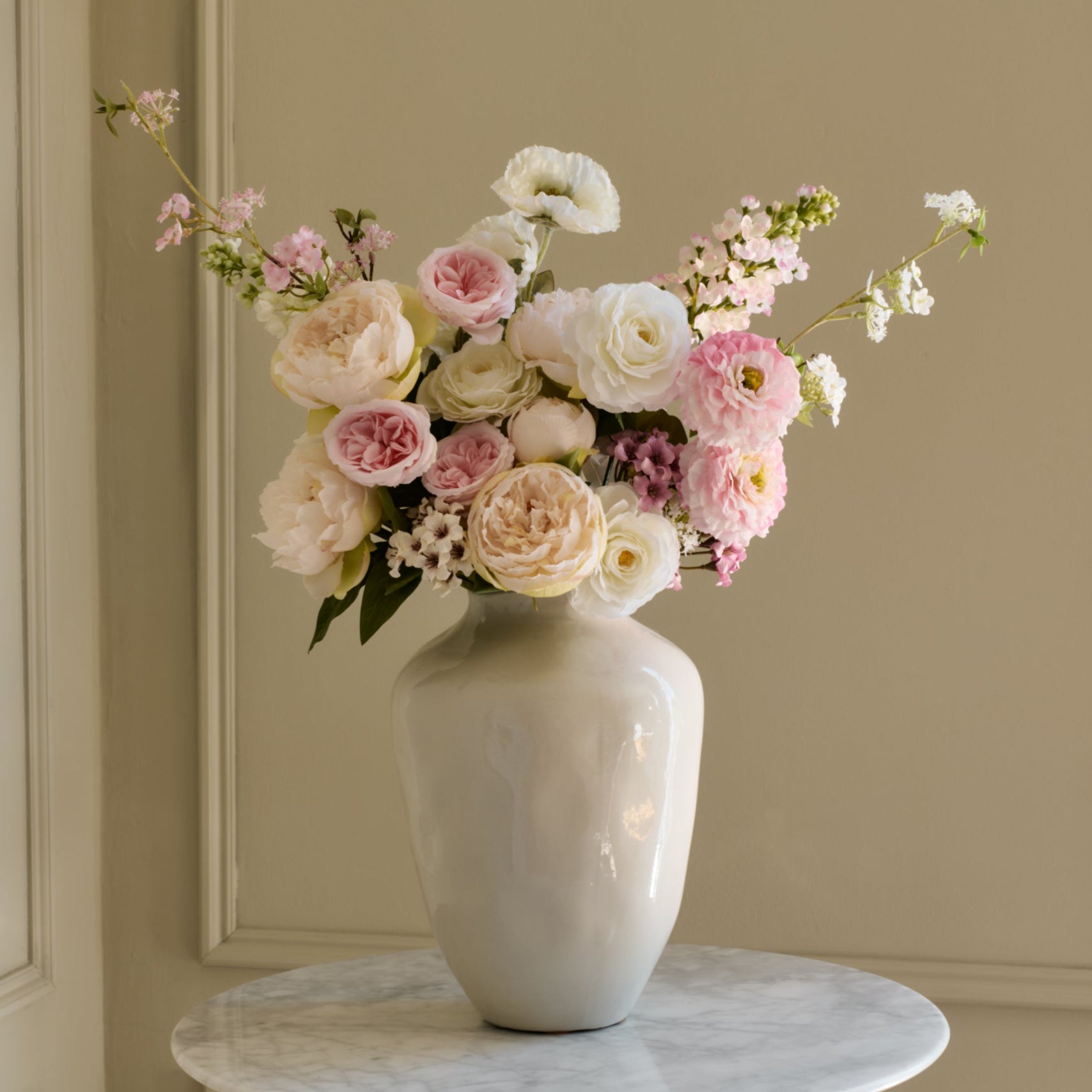 White vase with a bouquet of flowers on a marble surface against a beige wall.
