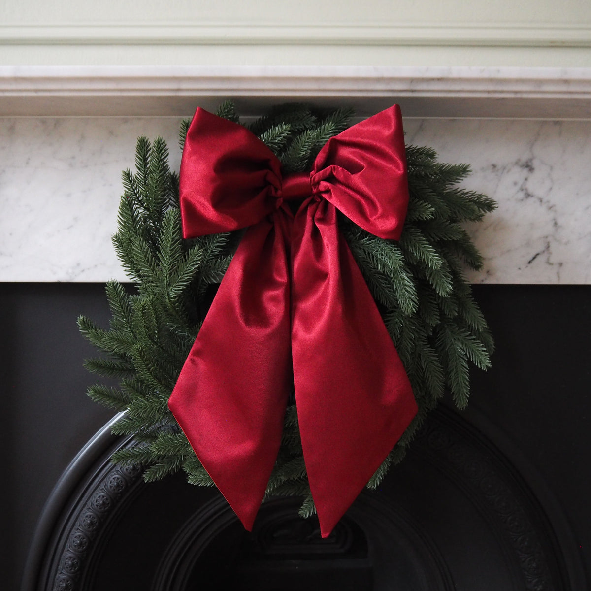 A green Christmas wreath with a red satin bow, displayed on a dark circular object, possibly a door or mantel.