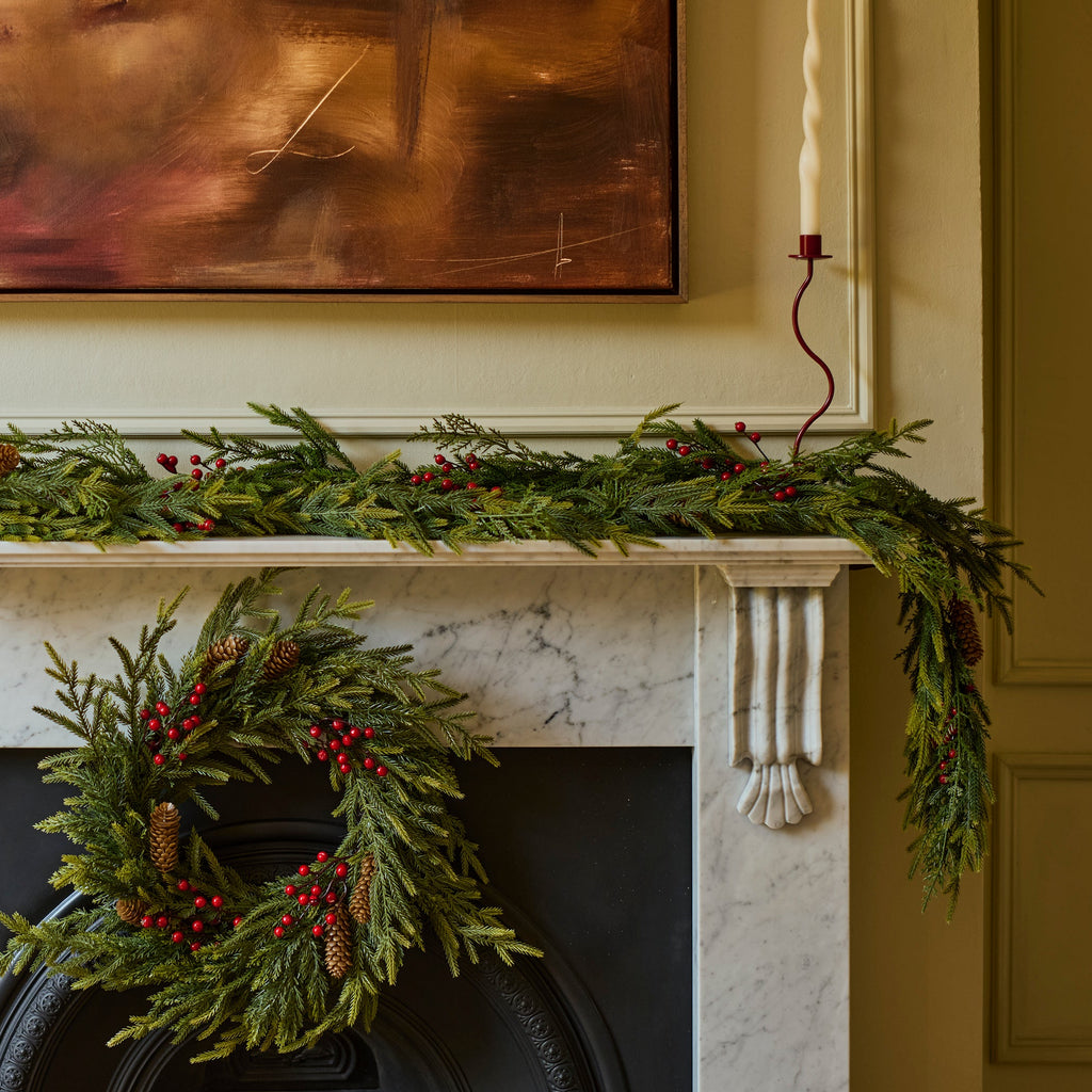 A holiday wreath made of green pine branches and red berries hangs on a black fireplace surround, with a matching green garland draped across a white mantel. A framed abstract painting and a candle are visible in the room.