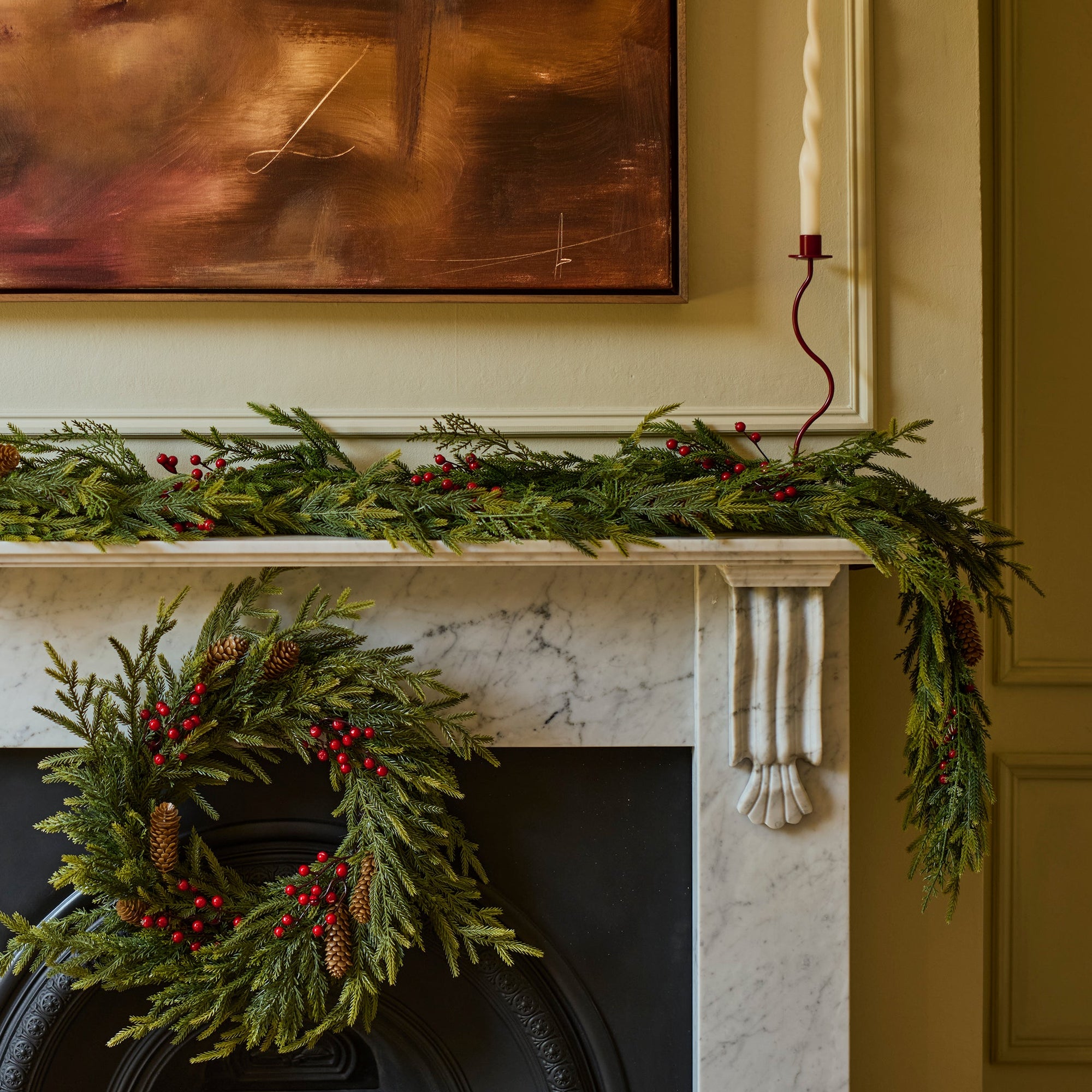 A holiday wreath made of green pine branches and red berries hangs on a black fireplace surround, with a matching green garland draped across a white mantel. A framed abstract painting and a candle are visible in the room.