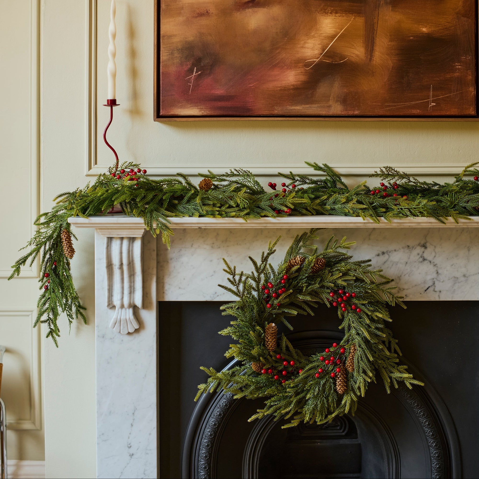 A holiday wreath made of green pine branches and red berries hangs on a black fireplace surround, with a matching green garland draped across a white mantel. A framed abstract painting and a candle are visible in the room.