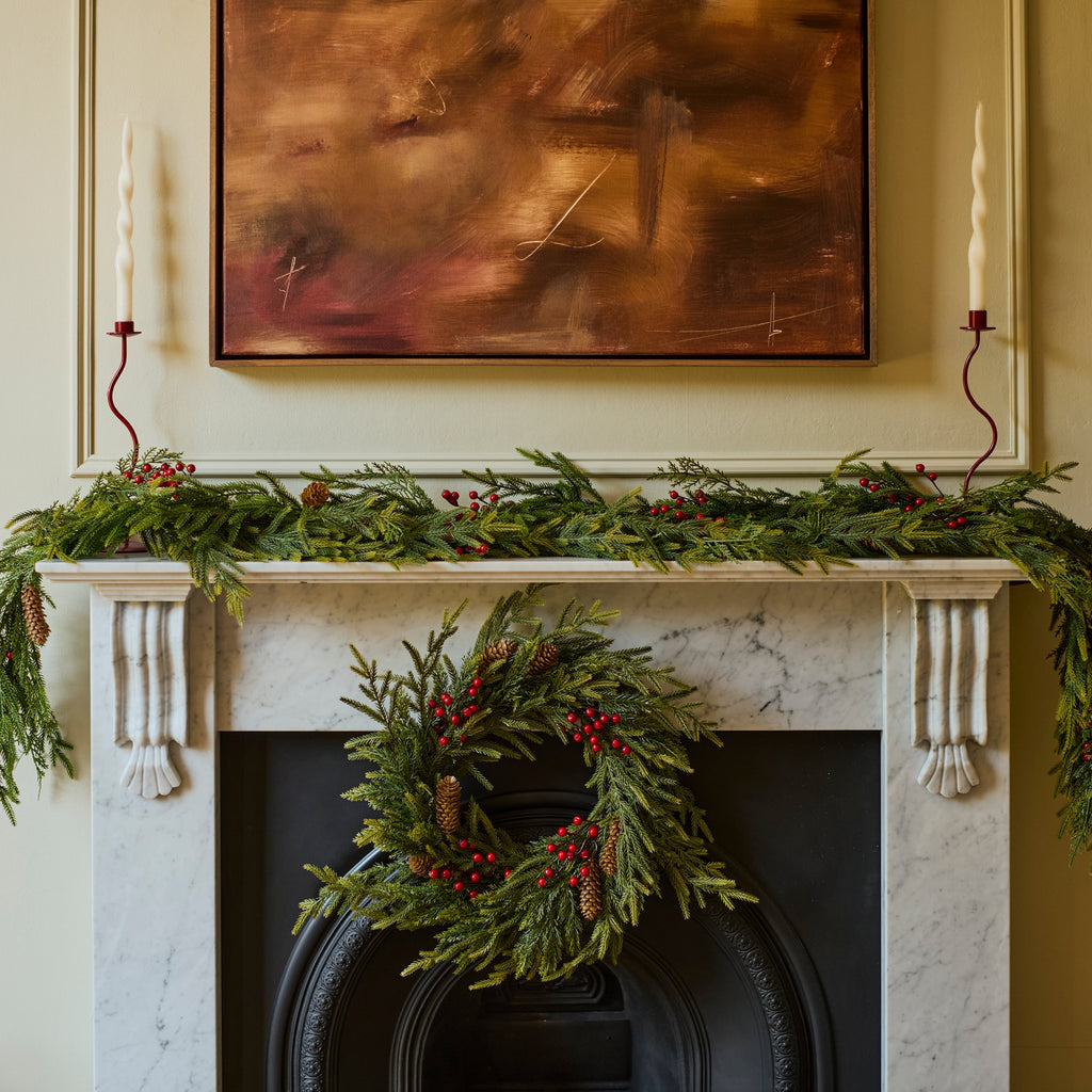 A holiday wreath made of green pine branches and red berries hangs on a black fireplace surround, with a matching green garland draped across a white mantel. A framed abstract painting and a candle are visible in the room.