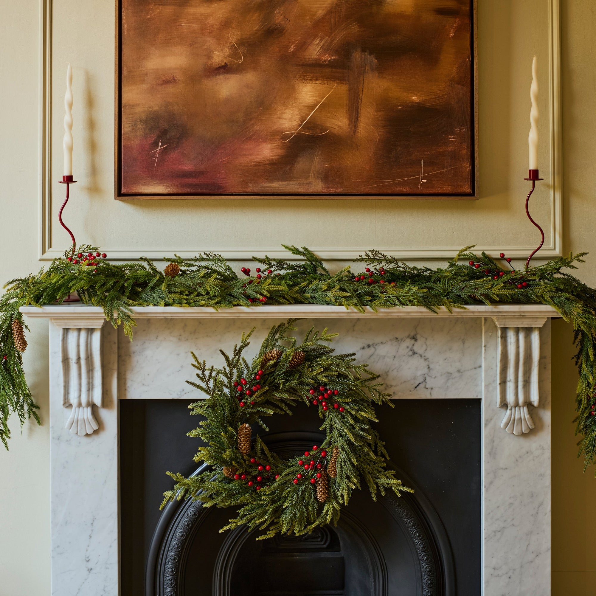 A holiday wreath made of green pine branches and red berries hangs on a black fireplace surround, with a matching green garland draped across a white mantel. A framed abstract painting and a candle are visible in the room.