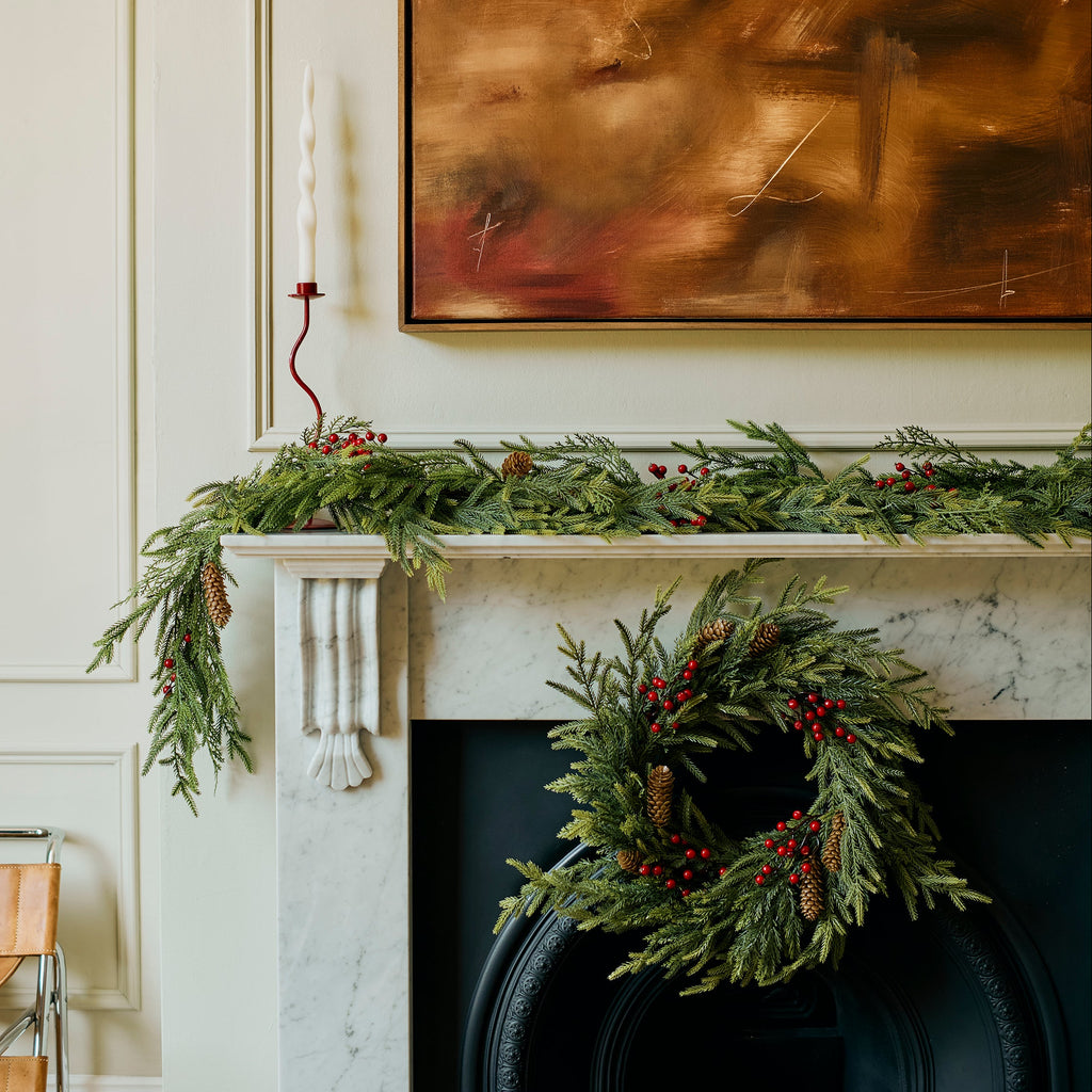 A holiday-themed interior scene featuring a white marble fireplace mantel decorated with a green garland and a matching green wreath with red berries, set against a cream paneled wall with a large abstract brown painting.