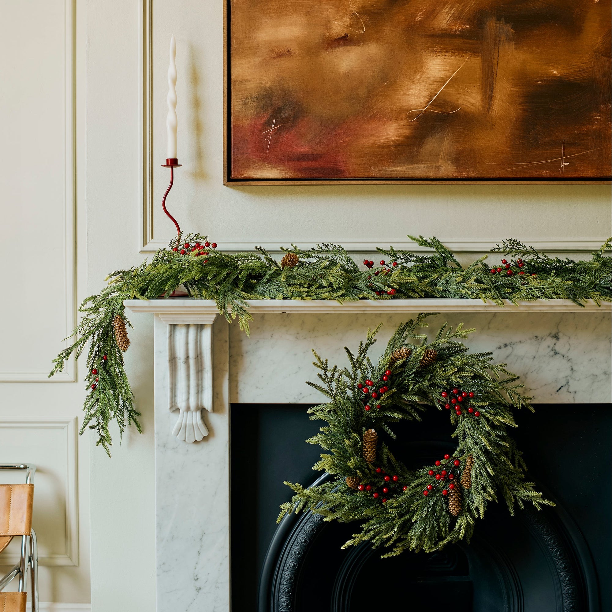 A holiday-themed interior scene featuring a white marble fireplace mantel decorated with a green garland and a matching green wreath with red berries, set against a cream paneled wall with a large abstract brown painting.