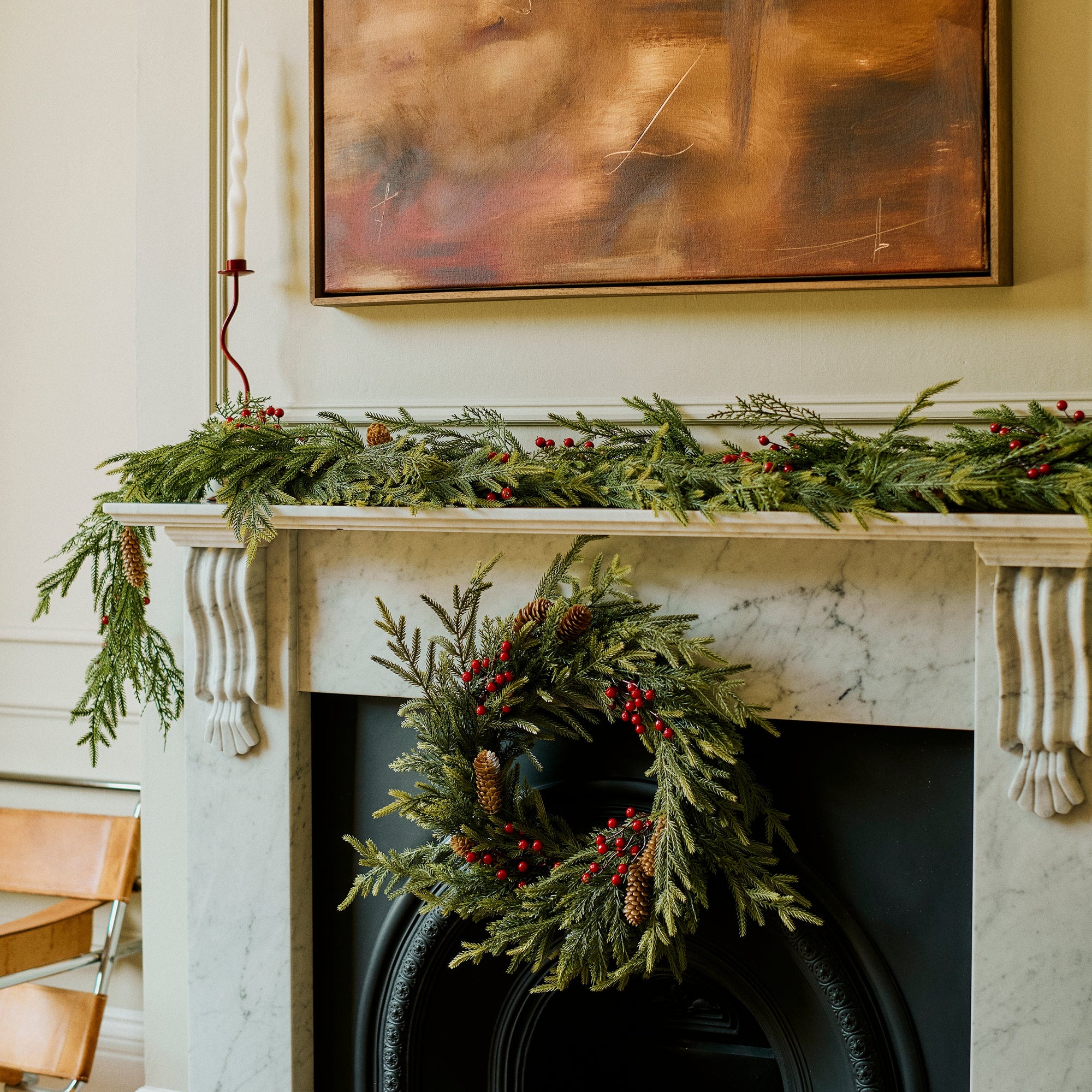 A festive holiday scene featuring a green garland and wreath decorated with red berries on a white marble fireplace mantel, with a large abstract painting hanging above.