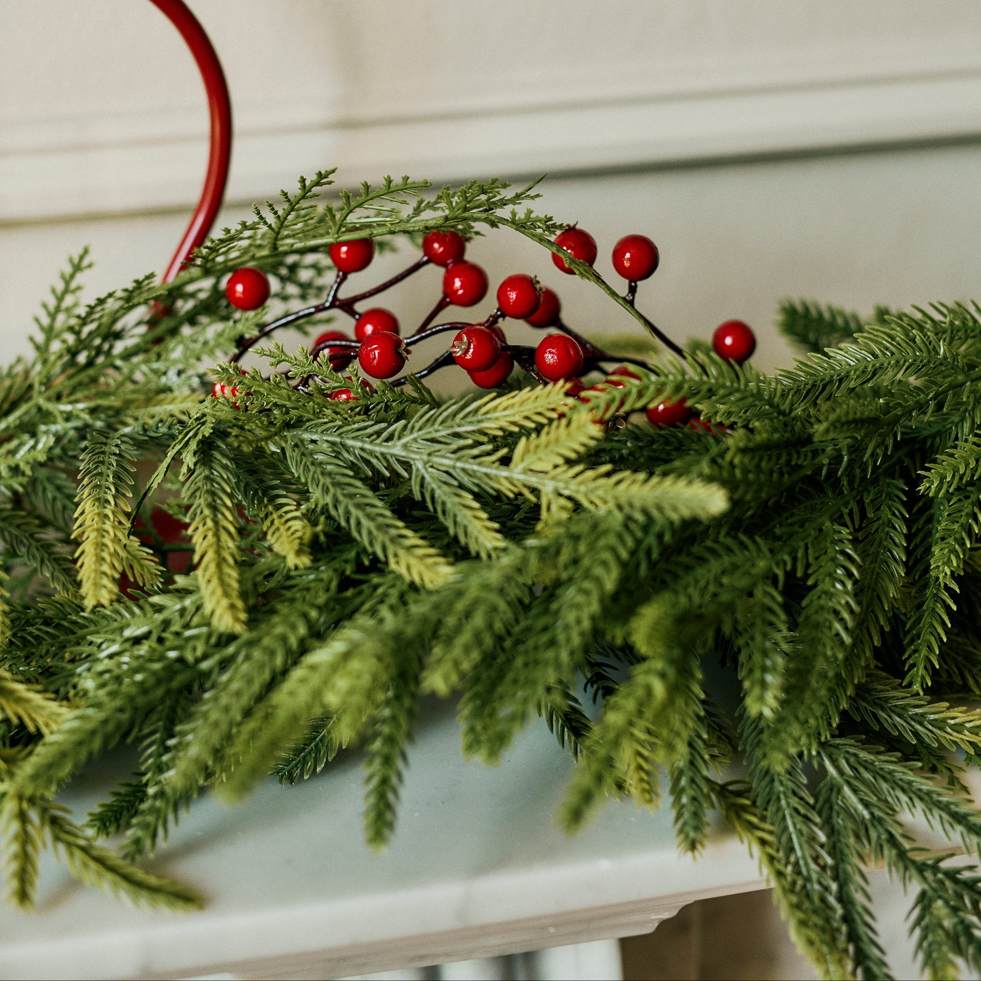 A green pine garland with clusters of red berries draped across a white mantel, with a red candlestick visible in the background against a cream wall.