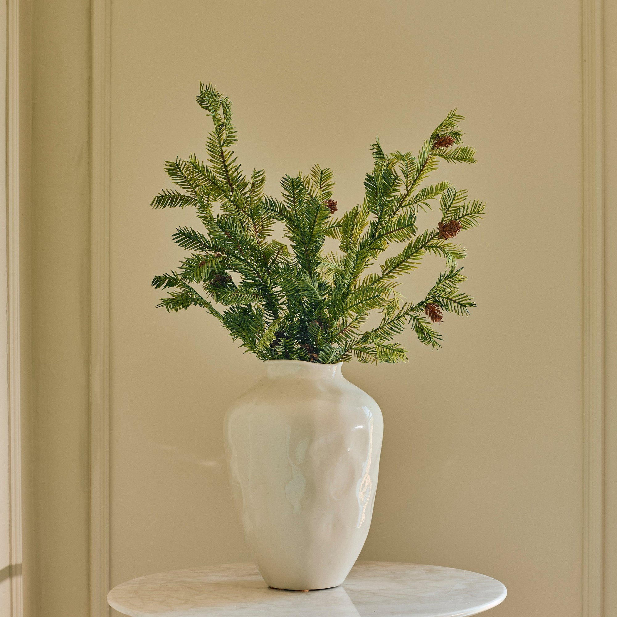 A green pine branch arrangement in a textured off-white ceramic vase, displayed on a small round white side table against a beige paneled wall.