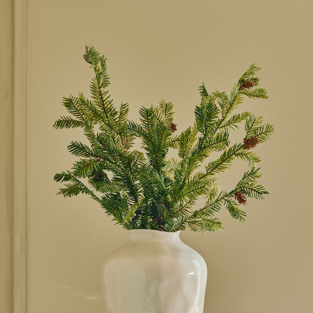 A green pine branch arrangement in a textured off-white ceramic vase, displayed on a small round white side table against a beige paneled wall.