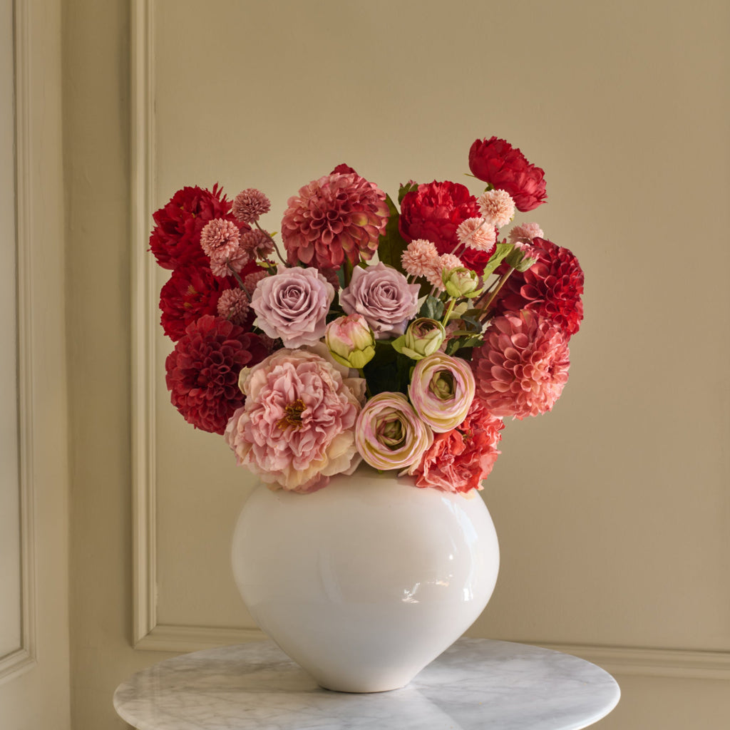 Bouquet of red, pink, and white flowers in a white vase on a marble surface with a beige wall background.