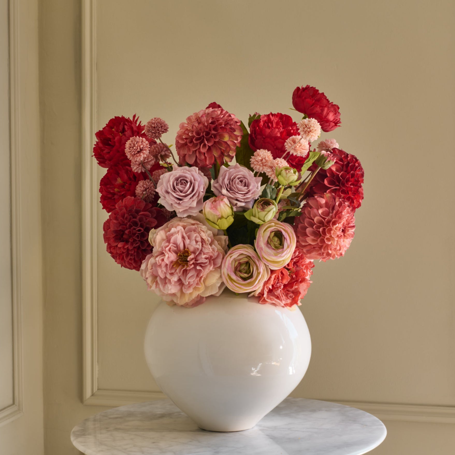 Bouquet of red, pink, and white flowers in a white vase on a marble surface with a beige wall background.