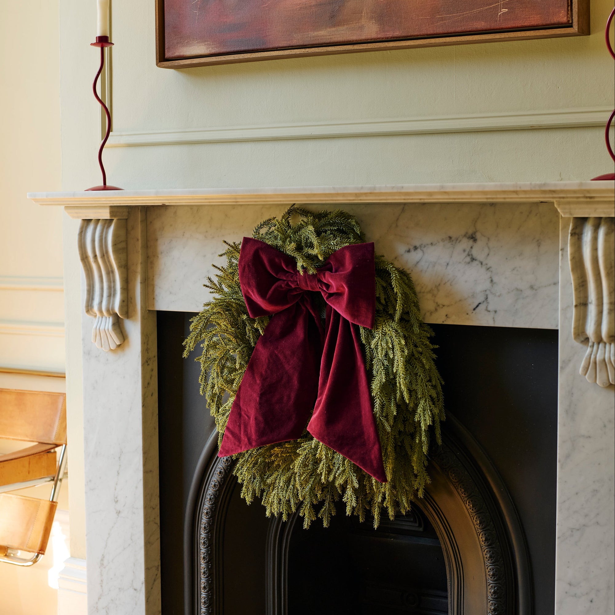 A green evergreen wreath with a large deep red velvet bow hanging on a white marble fireplace mantel in a decorated home interior.