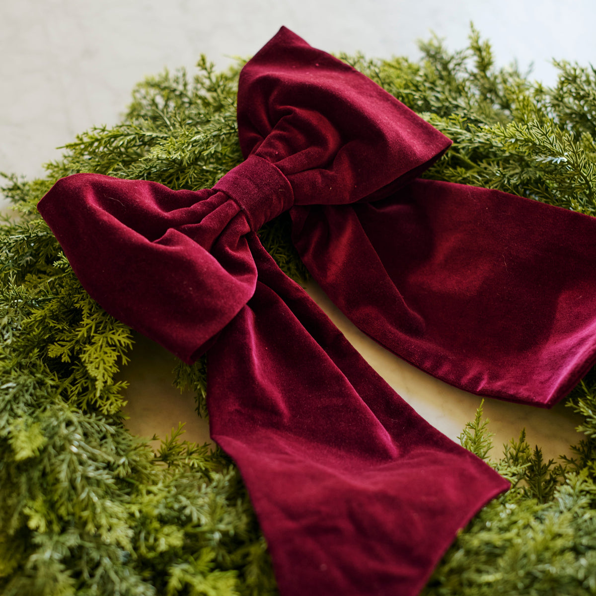 A large deep burgundy velvet bow tied on a green pine wreath, photographed against a light neutral background.