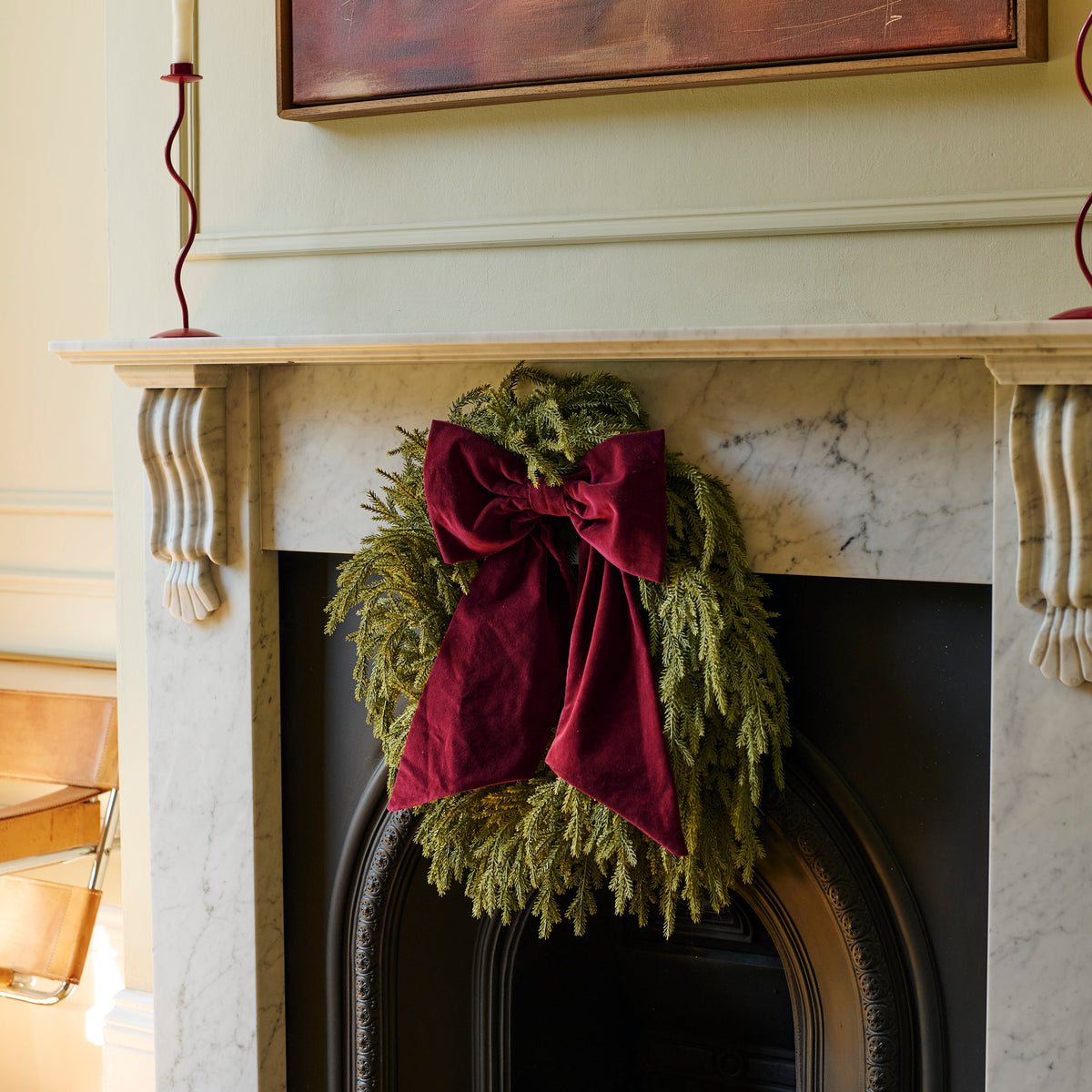 A green evergreen wreath with a large deep red velvet bow hanging on a white marble fireplace mantel in a decorated home interior.