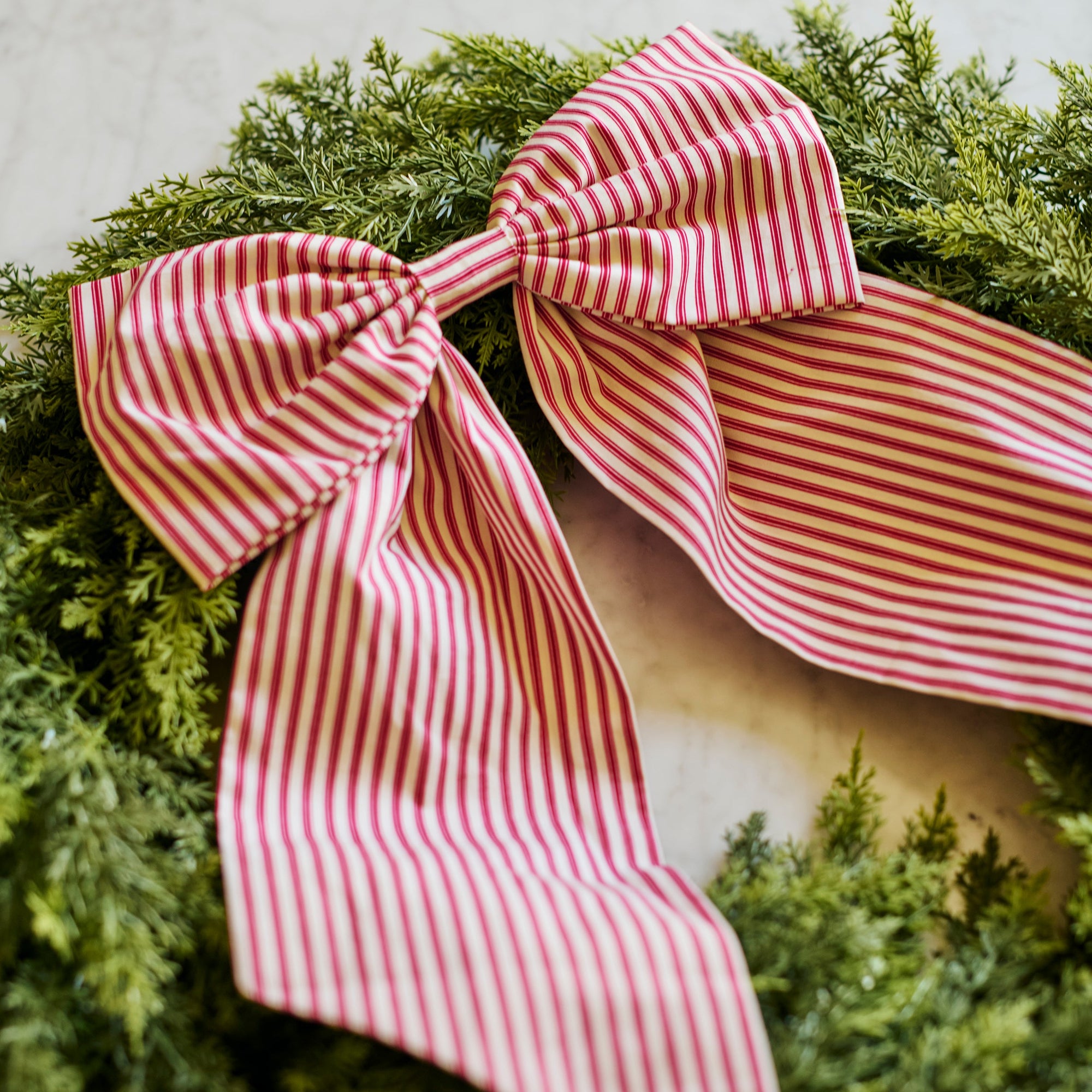 A large fabric bow with red and white vertical stripes tied in a knot, displayed on a green evergreen wreath against a light beige background.