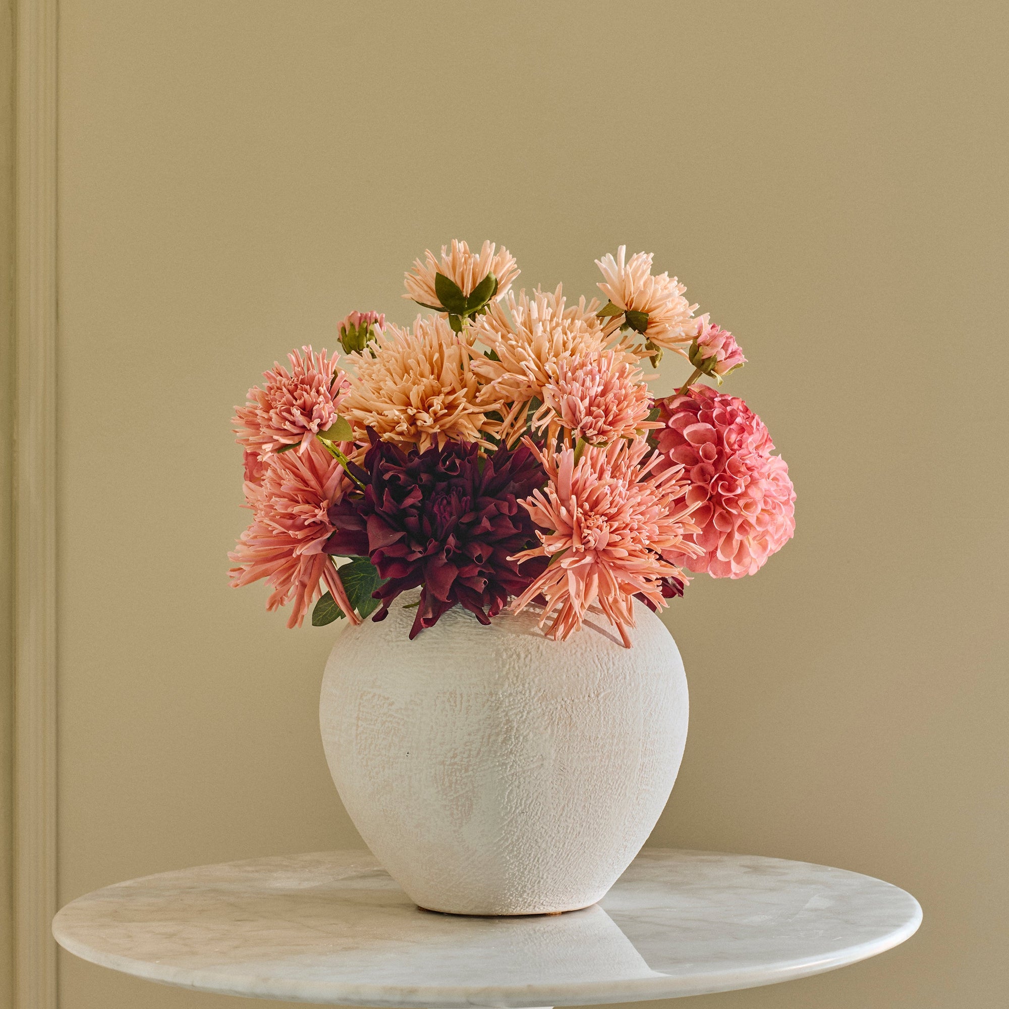 A white ceramic vase filled with pink, peach, and purple flowers, displayed on a small round white side table against a beige paneled wall.