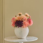 A white ceramic vase filled with pink, peach, and purple flowers, displayed on a small round white side table against a beige paneled wall.