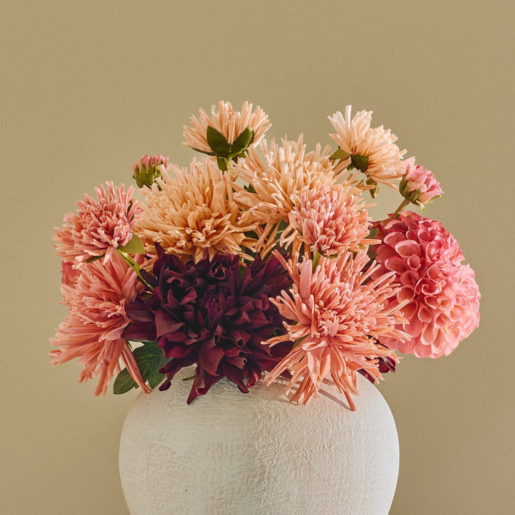 A white ceramic vase filled with pink, peach, and purple flowers, displayed on a small round white side table against a beige paneled wall.
