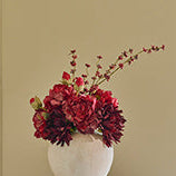A white ceramic vase filled with deep burgundy and red flowers, displayed on a small round white table against a beige paneled wall.