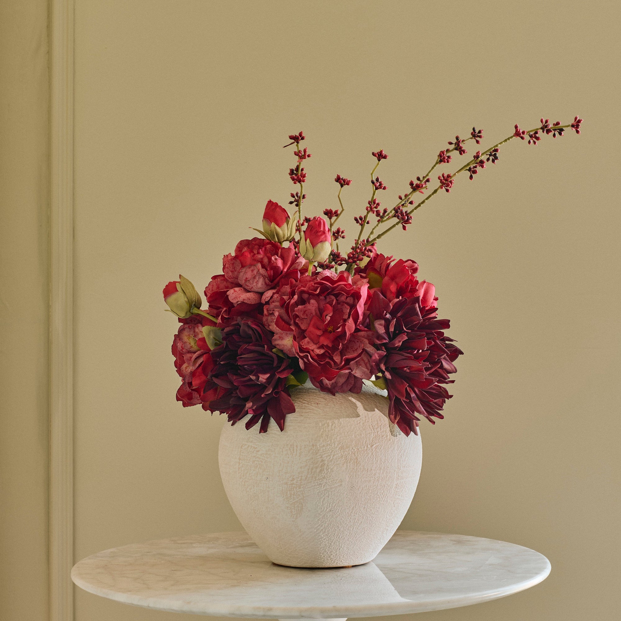 A textured off-white ceramic vase filled with deep burgundy and red floral arrangements, displayed on a round white marble side table against a beige paneled wall.