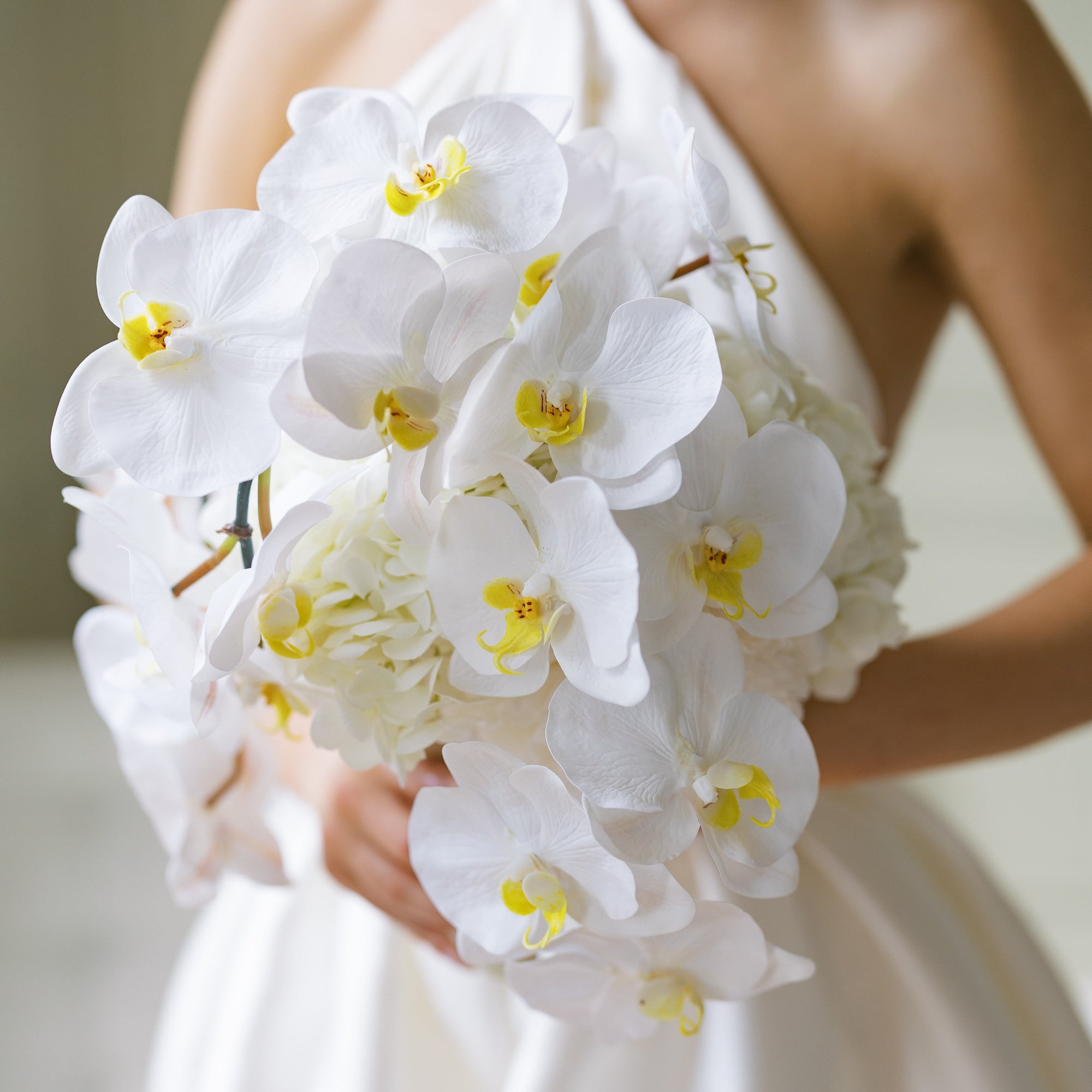 A bridal bouquet consisting of white peonies, hydrangeas, and orchids, tied with ribbon and held by a person in a white dress.