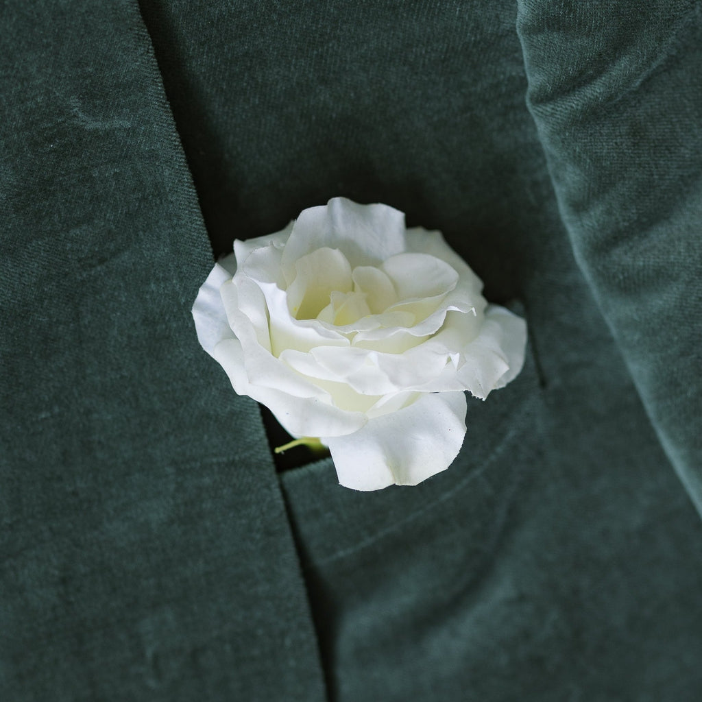 A close-up image of a white buttonhole flower attached to a dark green jacket, representing a real touch floral accessory for weddings.