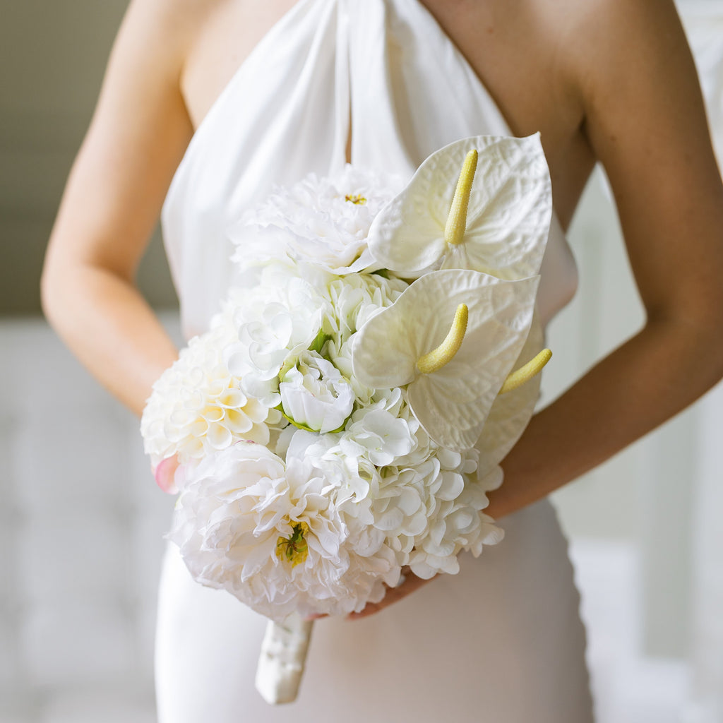 A bridal bouquet featuring white peonies, white hydrangeas, cream anthuriums, and a cream pom-pom, tied with white ribbon and held by a woman in a white dress.