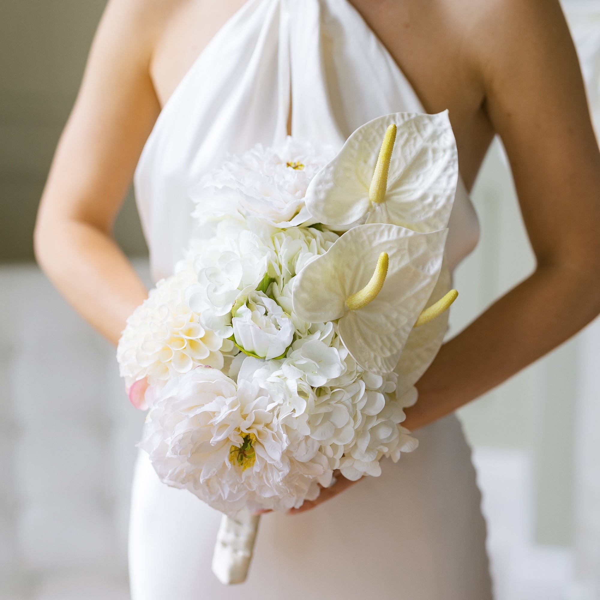 A bridal bouquet featuring white peonies, white hydrangeas, cream anthuriums, and a cream pom-pom, tied with white ribbon and held by a woman in a white dress.