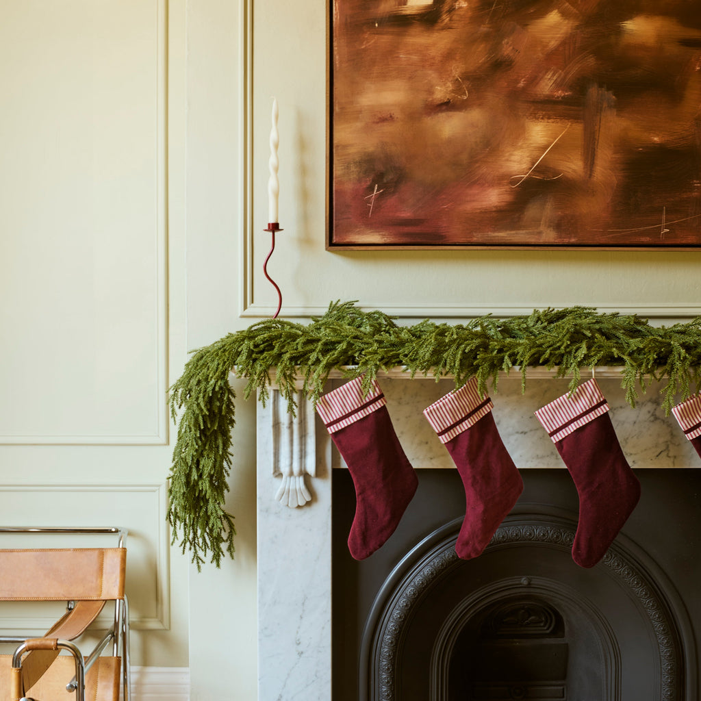 A styled holiday interior scene featuring a fireplace with a green pine garland and four burgundy Christmas stockings hanging from the mantel, beneath a large abstract painting.