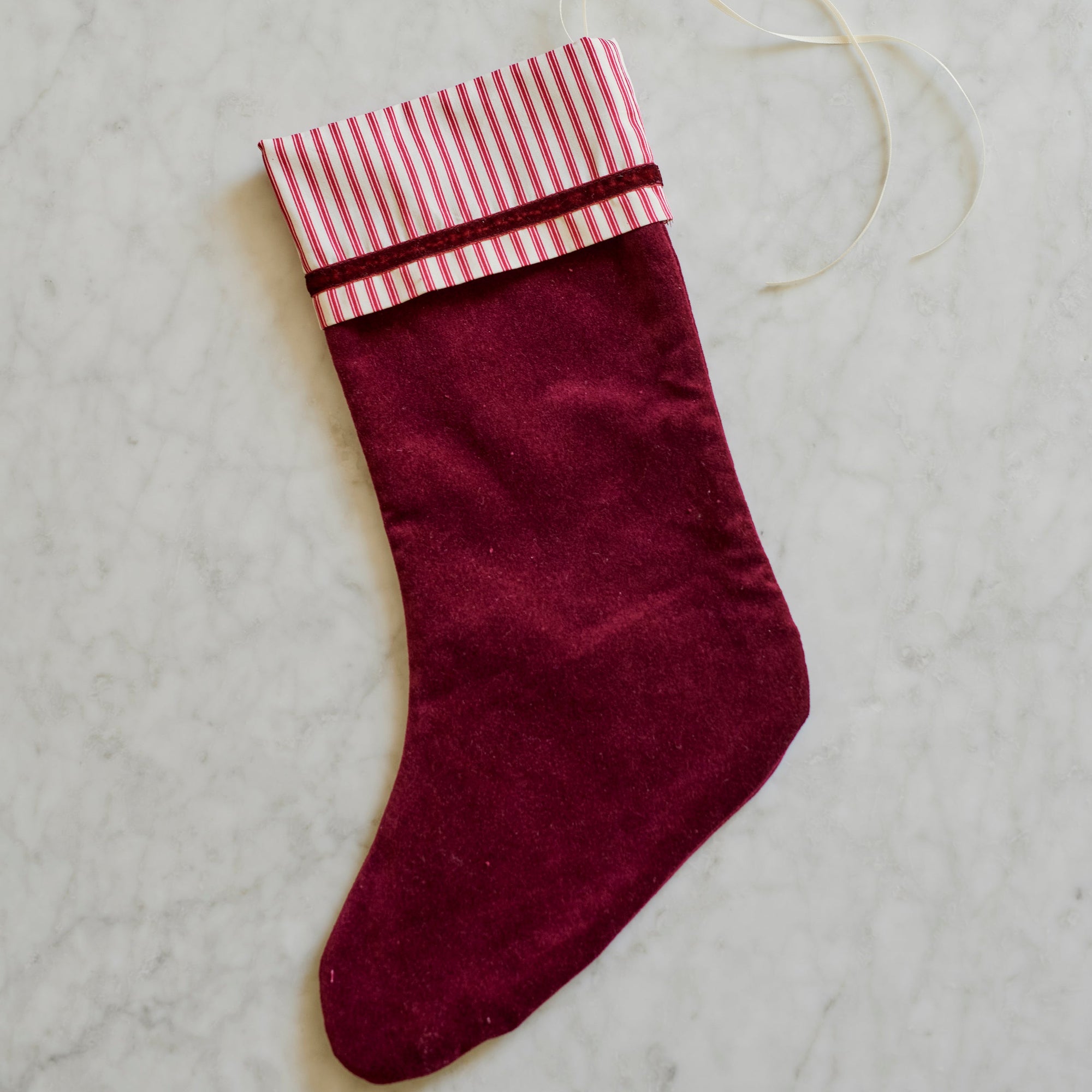 A burgundy velvet Christmas stocking with a pink and white striped cuff and a white hanging loop, shown on a light gray marble surface.
