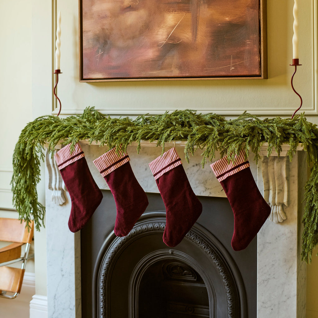 Four burgundy velvet Christmas stockings with pink striped cuffs hang from a green pine garland draped over a white marble fireplace mantel. A large abstract brown painting is mounted on the wall above the fireplace.