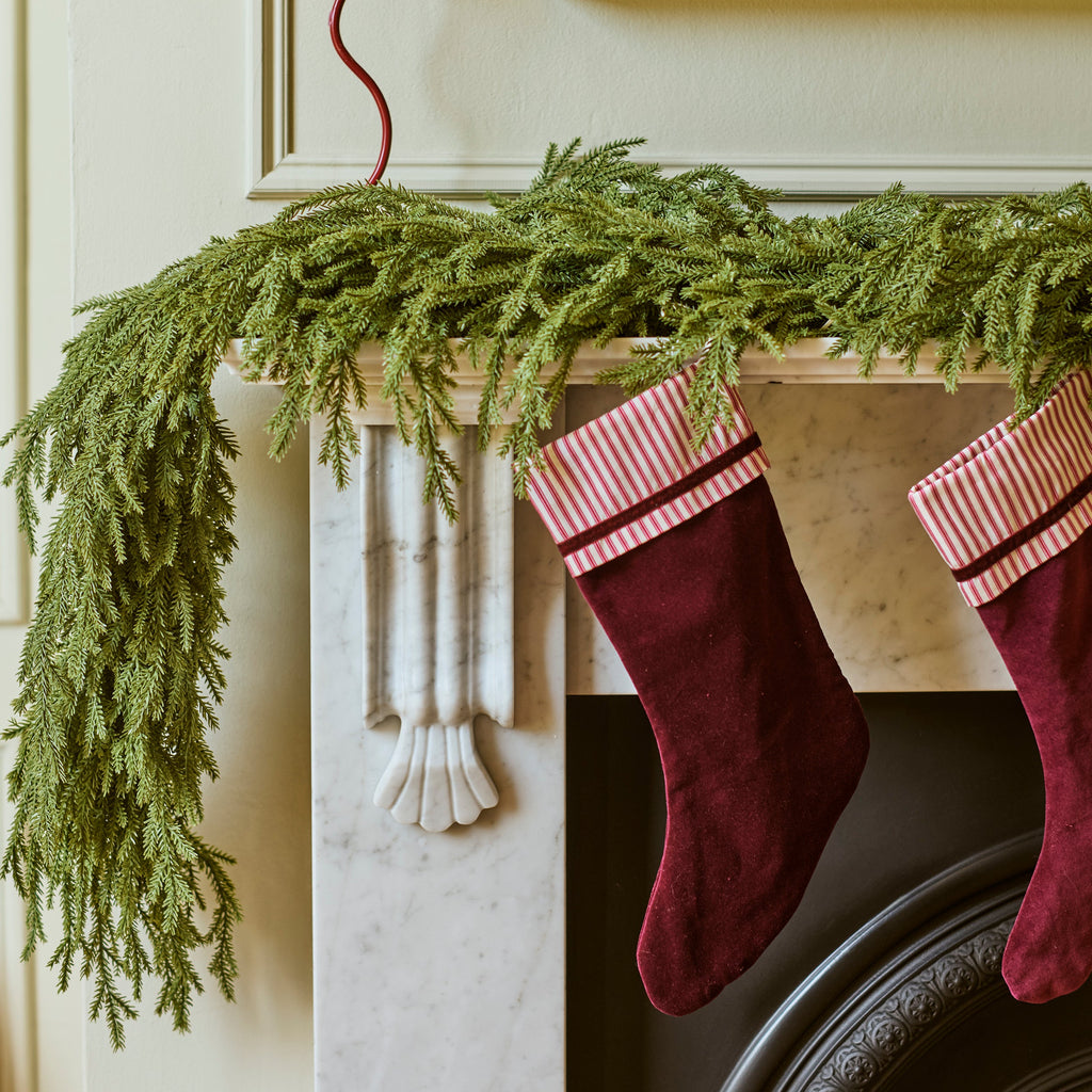 A holiday scene showing two burgundy velvet Christmas stockings with pink and white stripes hanging from a white marble fireplace mantel decorated with a green pine garland.