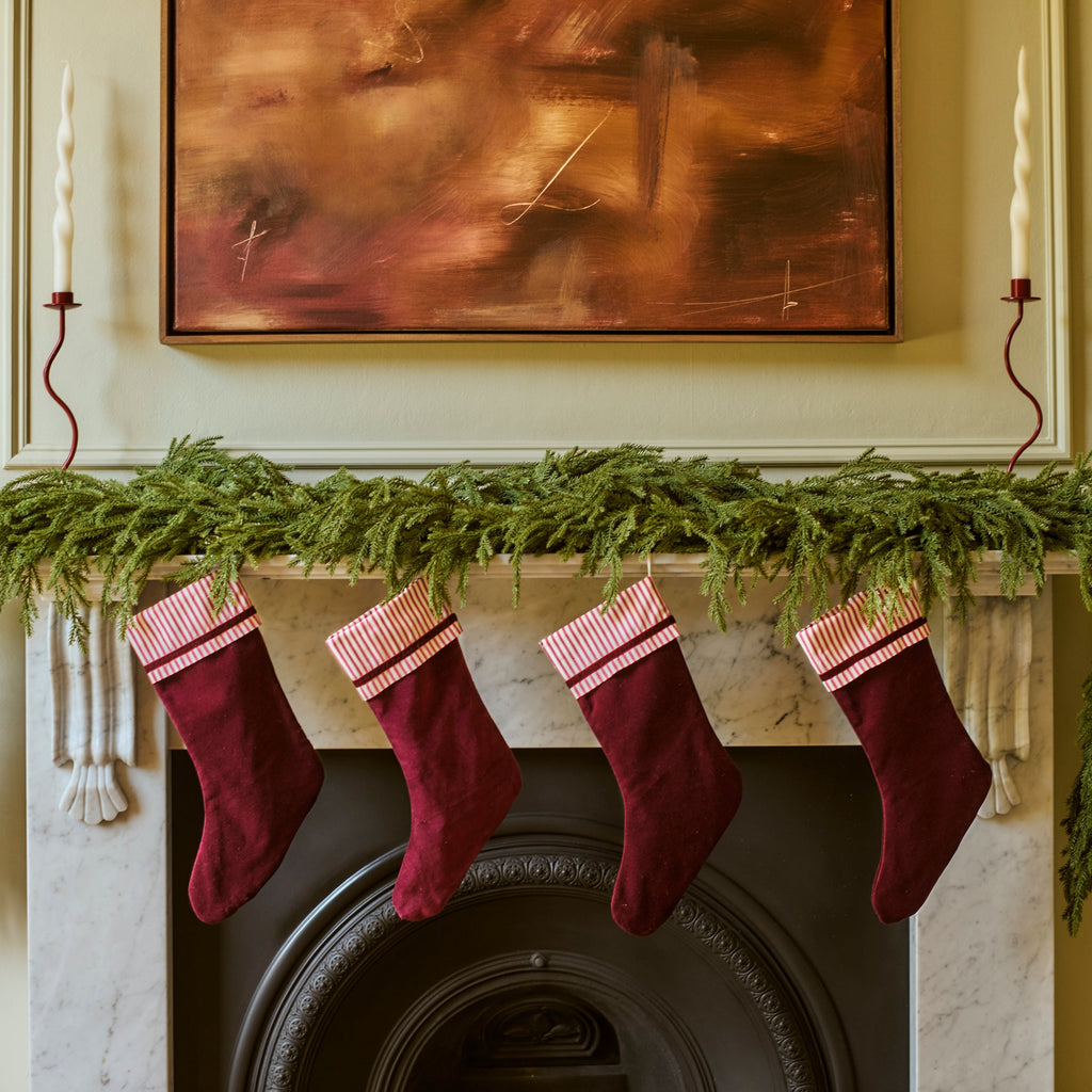 Four burgundy Christmas stockings with pink cuffs hang from a green garland on a fireplace mantel, with a framed painting above and white candles on either side.