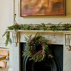 A festive holiday fireplace scene with a green pine garland draped across a white marble mantel and a matching wreath hanging in front of a black fireplace opening. A large framed abstract painting is mounted above the mantel.